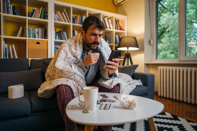 a man with a blanket draped over his shoulders drinks from a mug while viewing content on his smartphone.